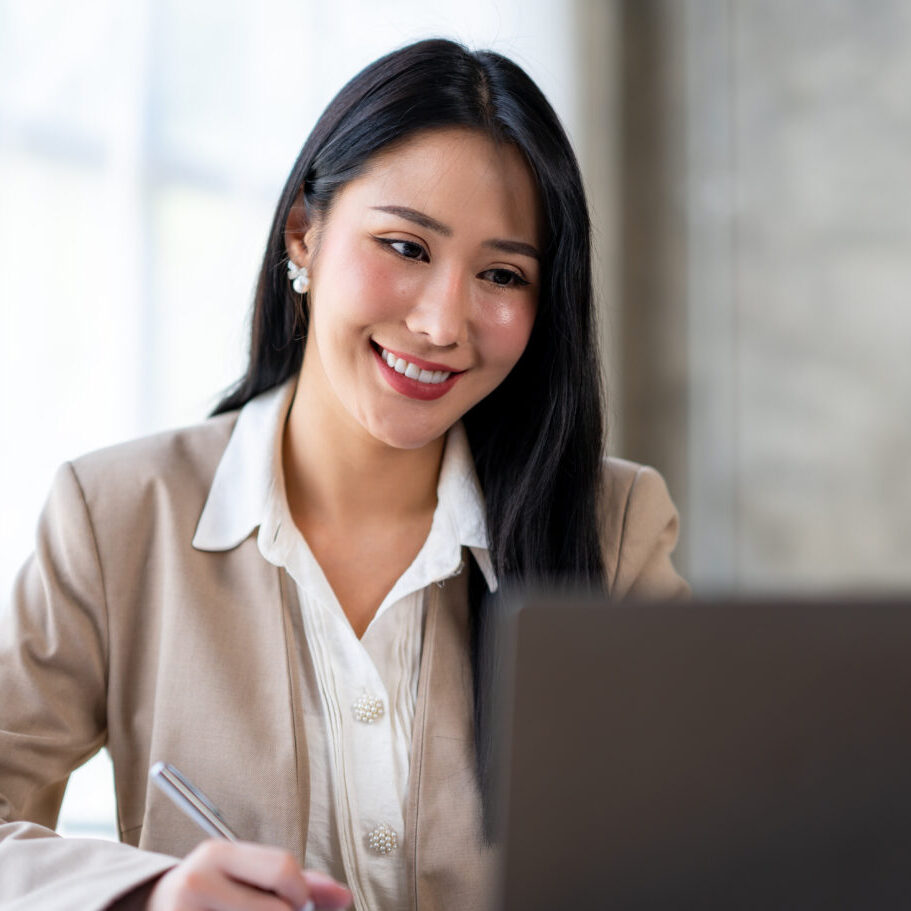 Confident Asian businesswoman sitting and taking notes in modern management concept with laptop computer in office happily.